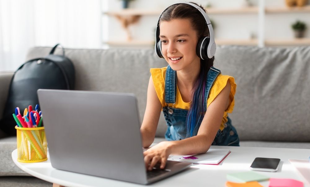Girl wearing headphones and denim overalls, using a laptop at a table, smiling.
