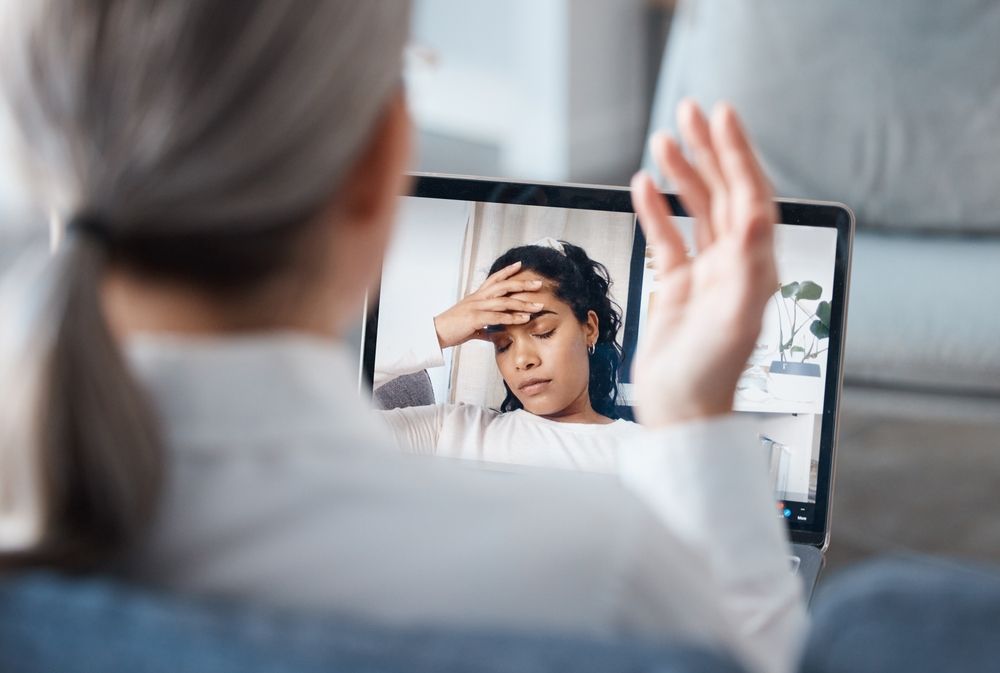 Person on laptop screen with hand on forehead. Another person gestures, likely in a telehealth session.