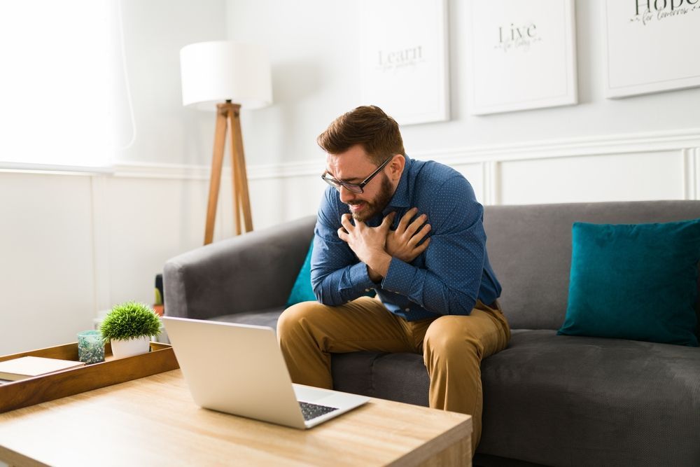 Man clutching chest, possibly in pain, seated on couch, looking at laptop in living room.