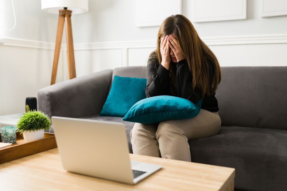 Woman sitting on a couch with hands covering face; laptop on table.