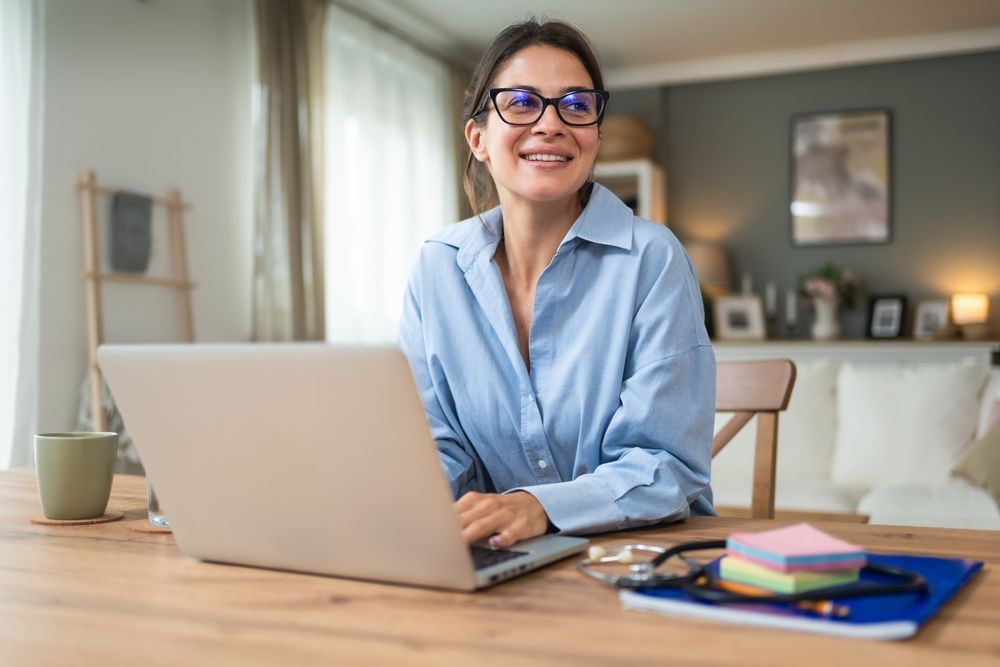 Woman with glasses smiling, working on a laptop at a wooden table in a well-lit room.