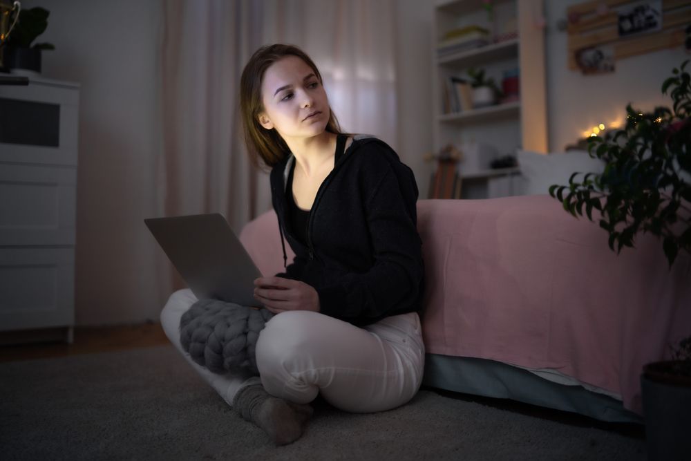Woman in a dark room with a laptop sits on the floor near a bed, looking off to the side.