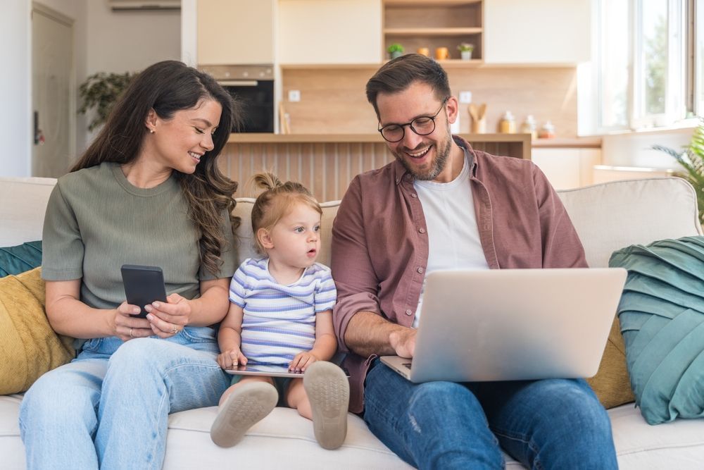Family of three on a sofa, using a phone, laptop, and tablet; home setting.