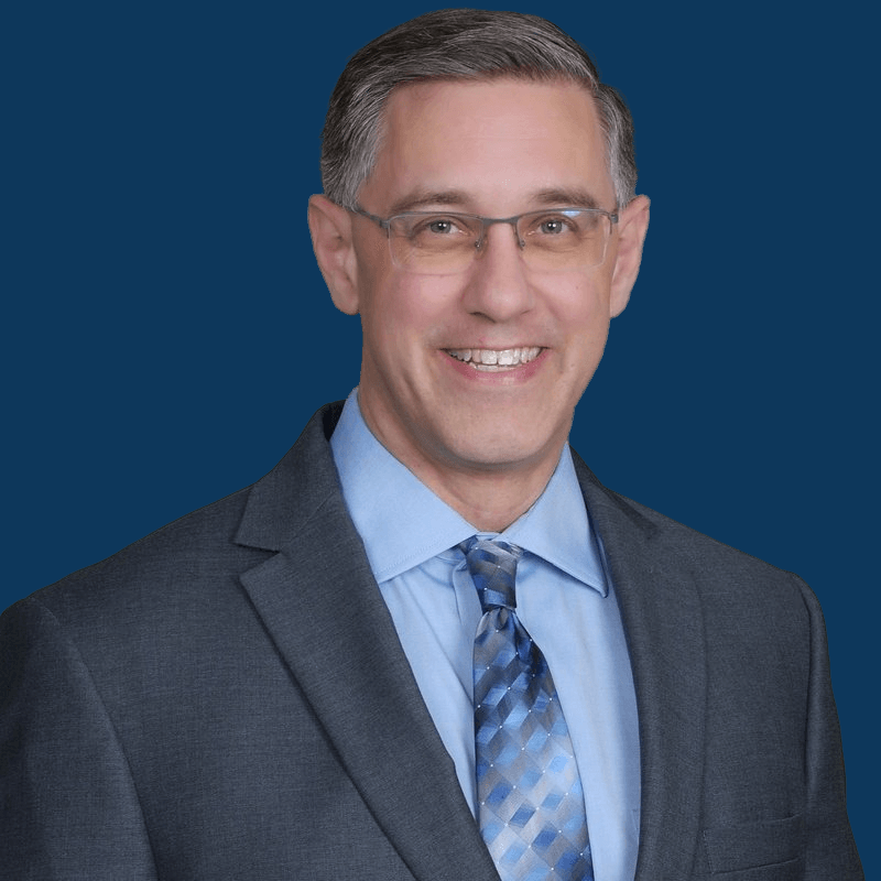 Headshot of a smiling person wearing glasses, a light blue collared shirt, a patterned blue tie, and a gray suit jacket.