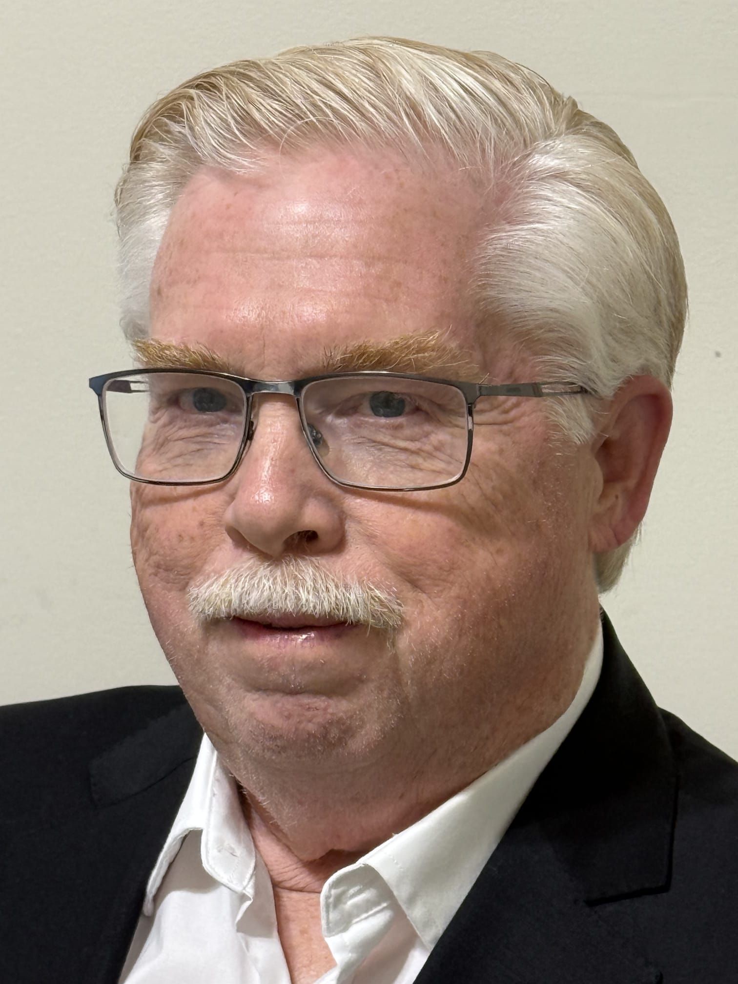 A person with white hair and a mustache wearing glasses, a white collared shirt, and a black blazer against a plain wall.
