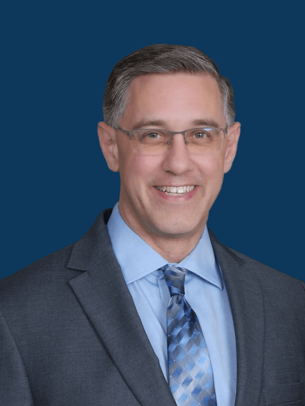 Man in suit smiles, indoors, possibly a business setting.