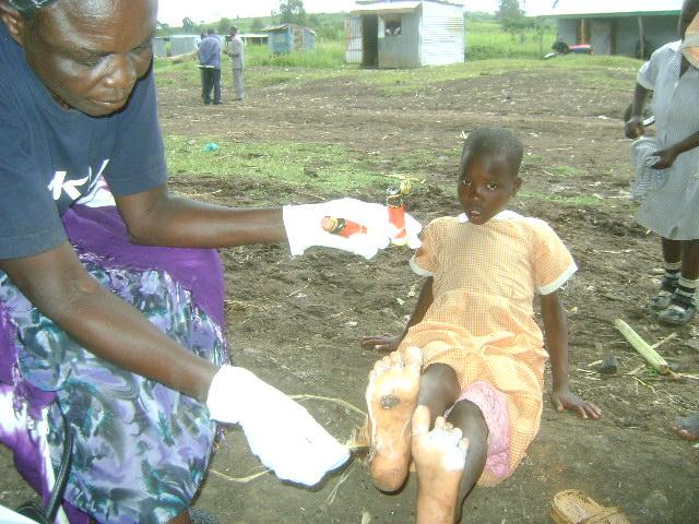 Person in gloves handing an item to a child seated outdoors on bare ground.