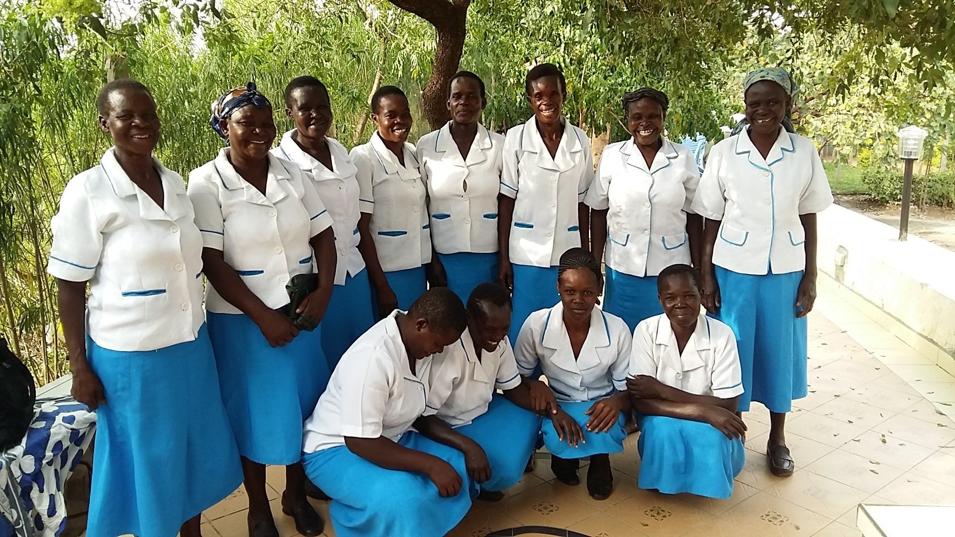 Group of students in white shirts and blue skirts posing outdoors under a tree