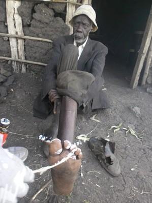 Person outdoors holding a large wooden pipe or tool, seated near stacked materials and a doorway.