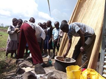 People gathering around a yellow water tank, filling buckets outdoors beside a thatched wall