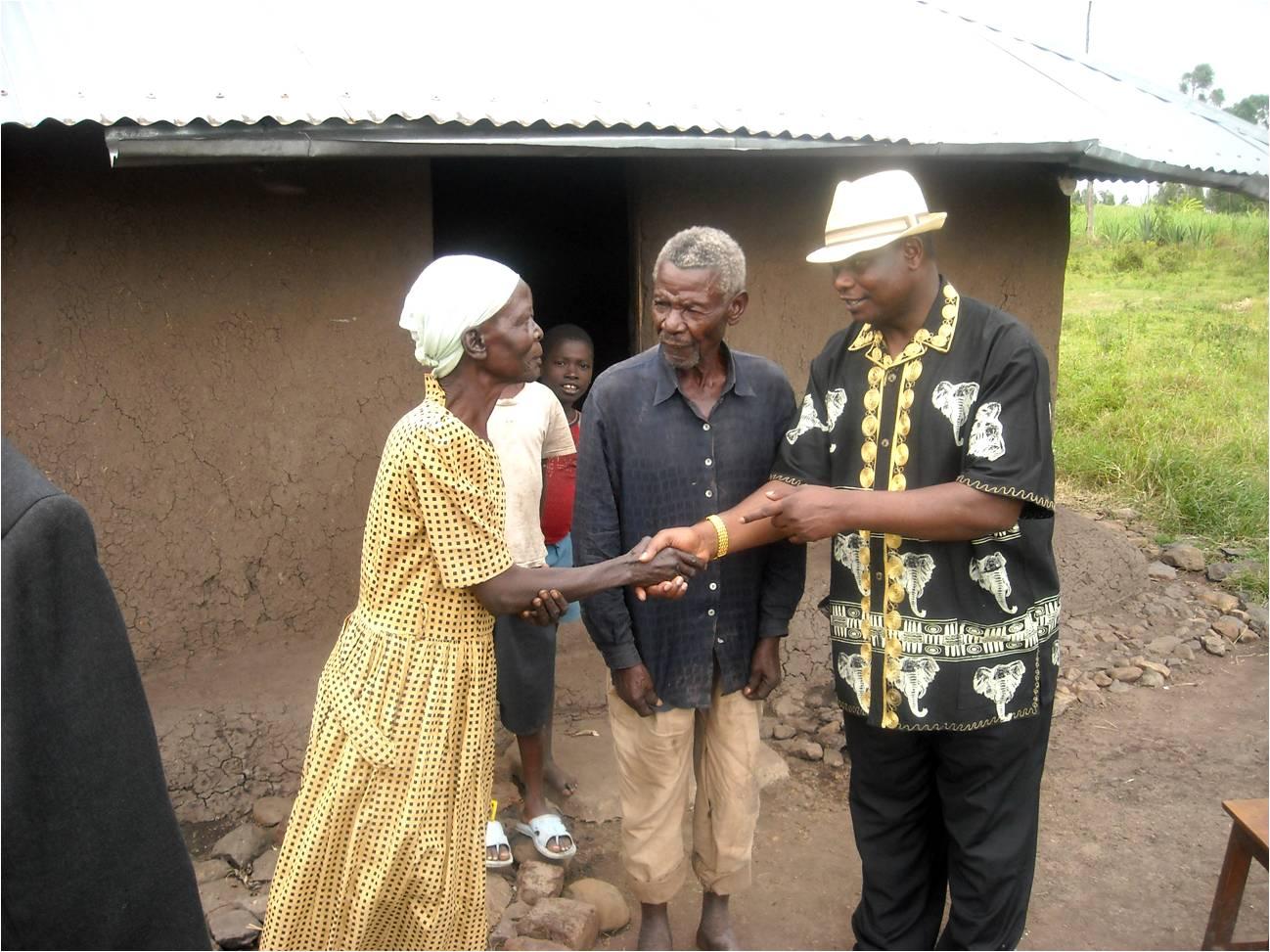 Four people stand outside a small building, shaking hands over a document or object.