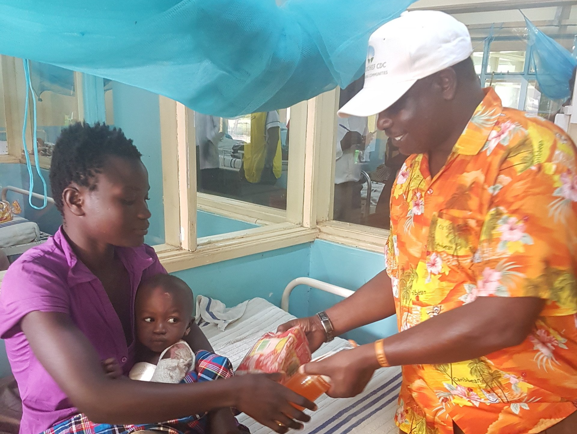 Nurse handing a wrapped baby to a seated woman in a hospital ward under a blue mosquito net.