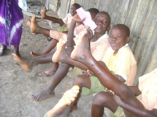 Children sitting outside a wooden building, smiling and holding up their feet toward the camera