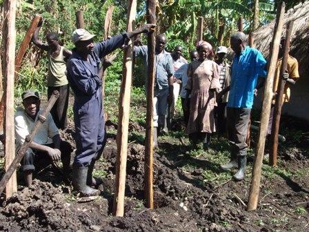 People planting seedlings in a muddy garden, with one person pointing and others standing among rows of stakes.