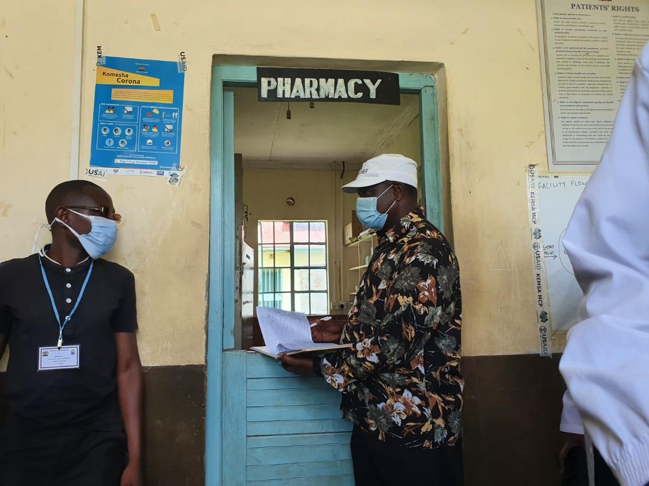 Two masked people standing at a pharmacy doorway, one holding papers.