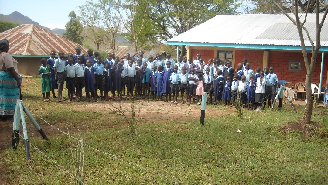 Crowd gathered outside a rural building with red walls and white roof, standing in a grassy yard.