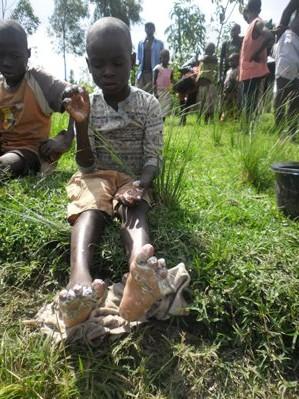 Child sitting on grass with muddy feet, holding a bottle, while others stand in the background.