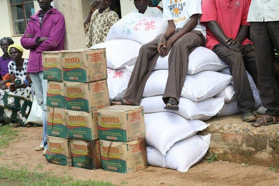 Stacked sacks and boxes of supplies beside seated people outdoors on a stone step.