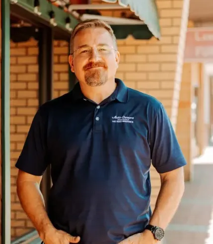 Man in a blue polo shirt with glasses, standing in front of a brick building, smiling.