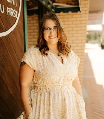 Woman with glasses, smiling in yellow dress, standing outside a building.