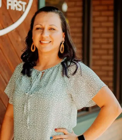 Woman in a green top with gold earrings smiles, hand on hip; in front of a building.