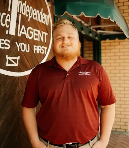 Man with red beard and polo shirt stands outside a building with a sign.
