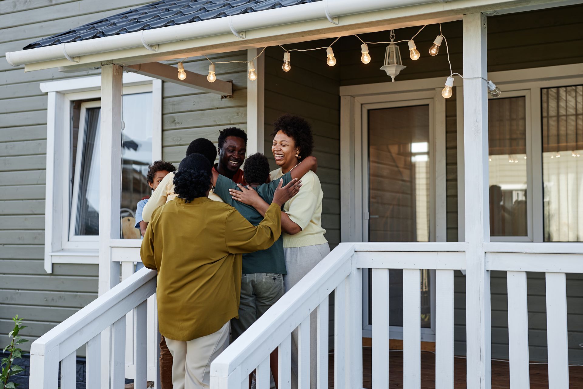 Family members embrace on a porch, lit by string lights; smiles, light beige paint, and greenery visible.