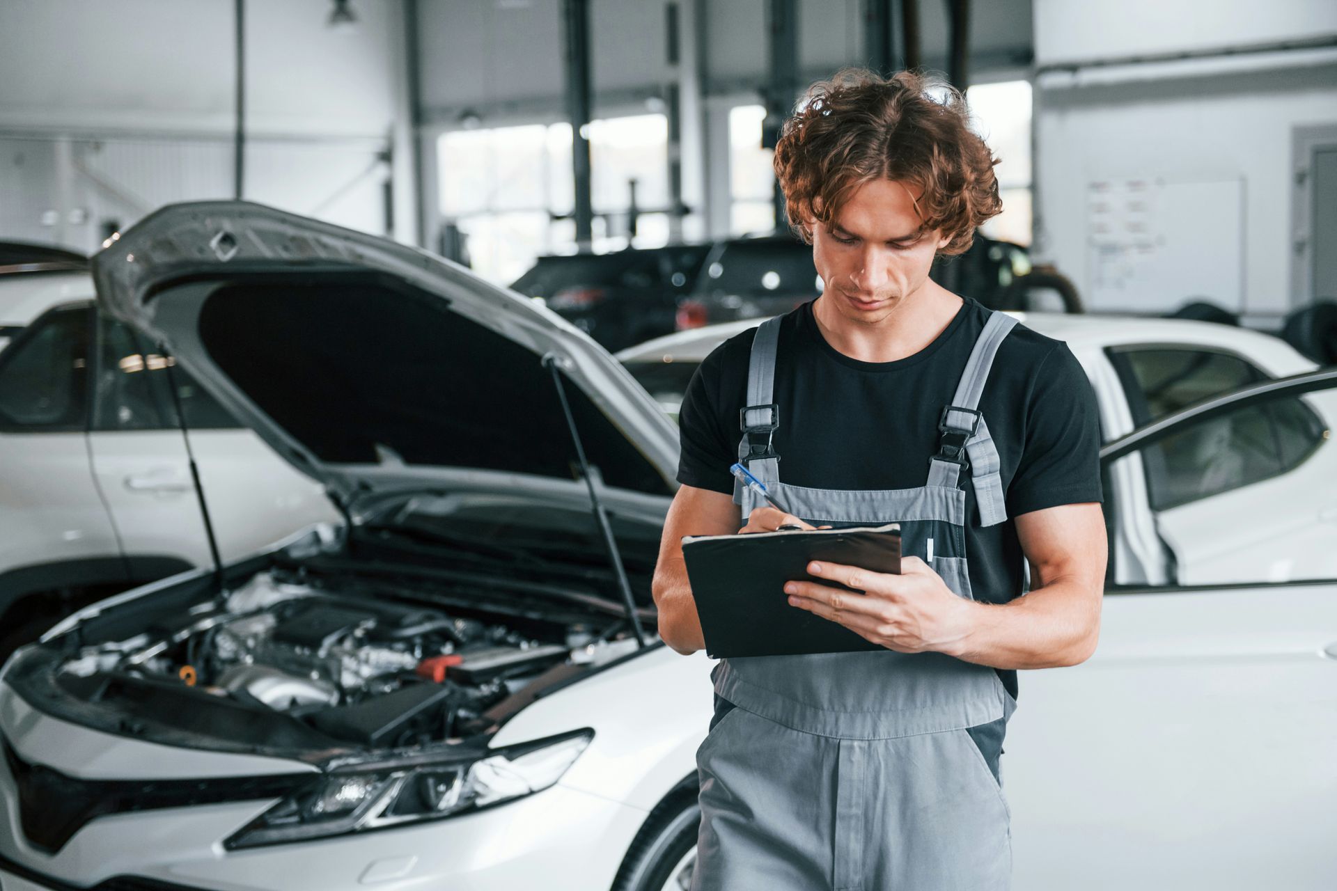 Mechanic in gray overalls inspects a car engine with the hood open, writing on a clipboard in a garage.