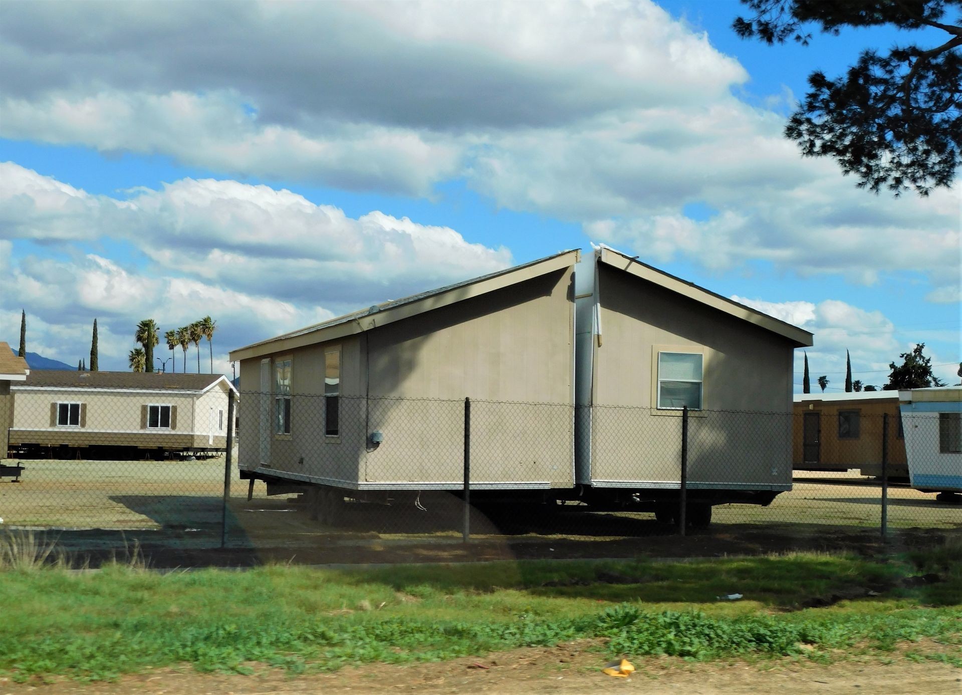 Mobile homes sit behind a chain-link fence under a cloudy blue sky. Green grass in the foreground.