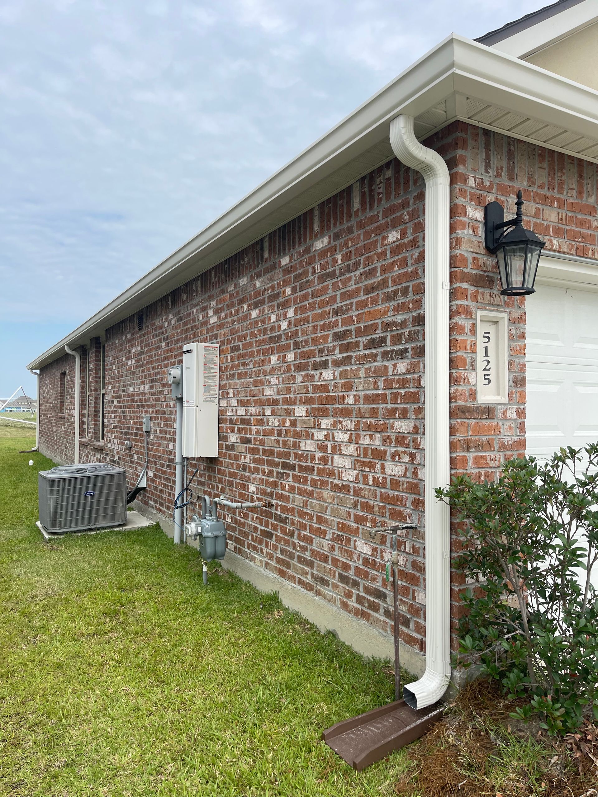 Red brick residential house exterior with white rain gutter downspout, air conditioning unit, and utility meter on the side wall.