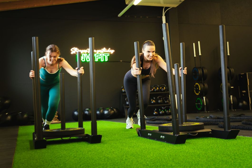 Two people pushing sleds on green turf in a gym, exercising under bright lights