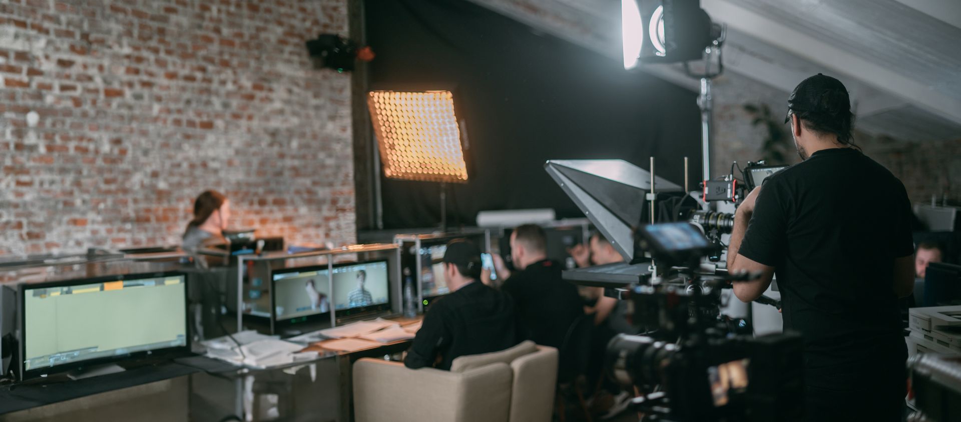 Film crew in a brick studio sets up cameras and monitors around a seated person.