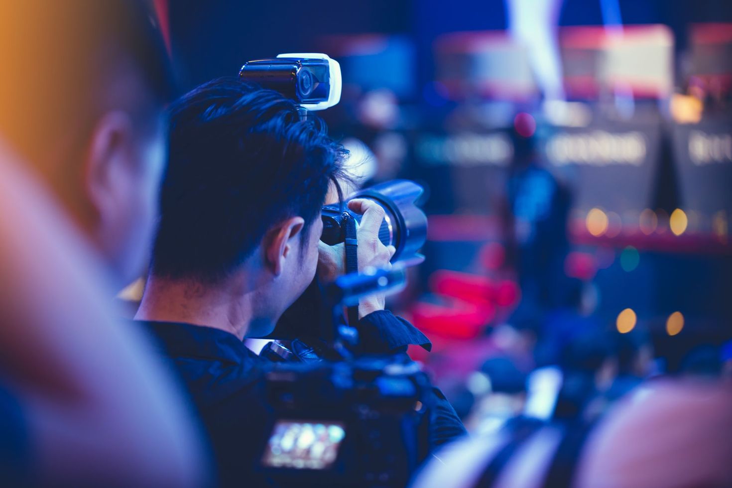 Camera operator filming at a brightly lit event with blurred audience lights in the background