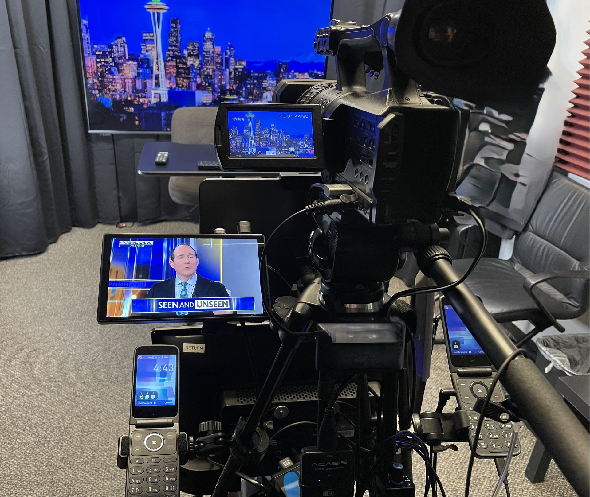 Video camera pointed at a home studio desk with multiple monitors showing a news broadcast and city skyline background.
