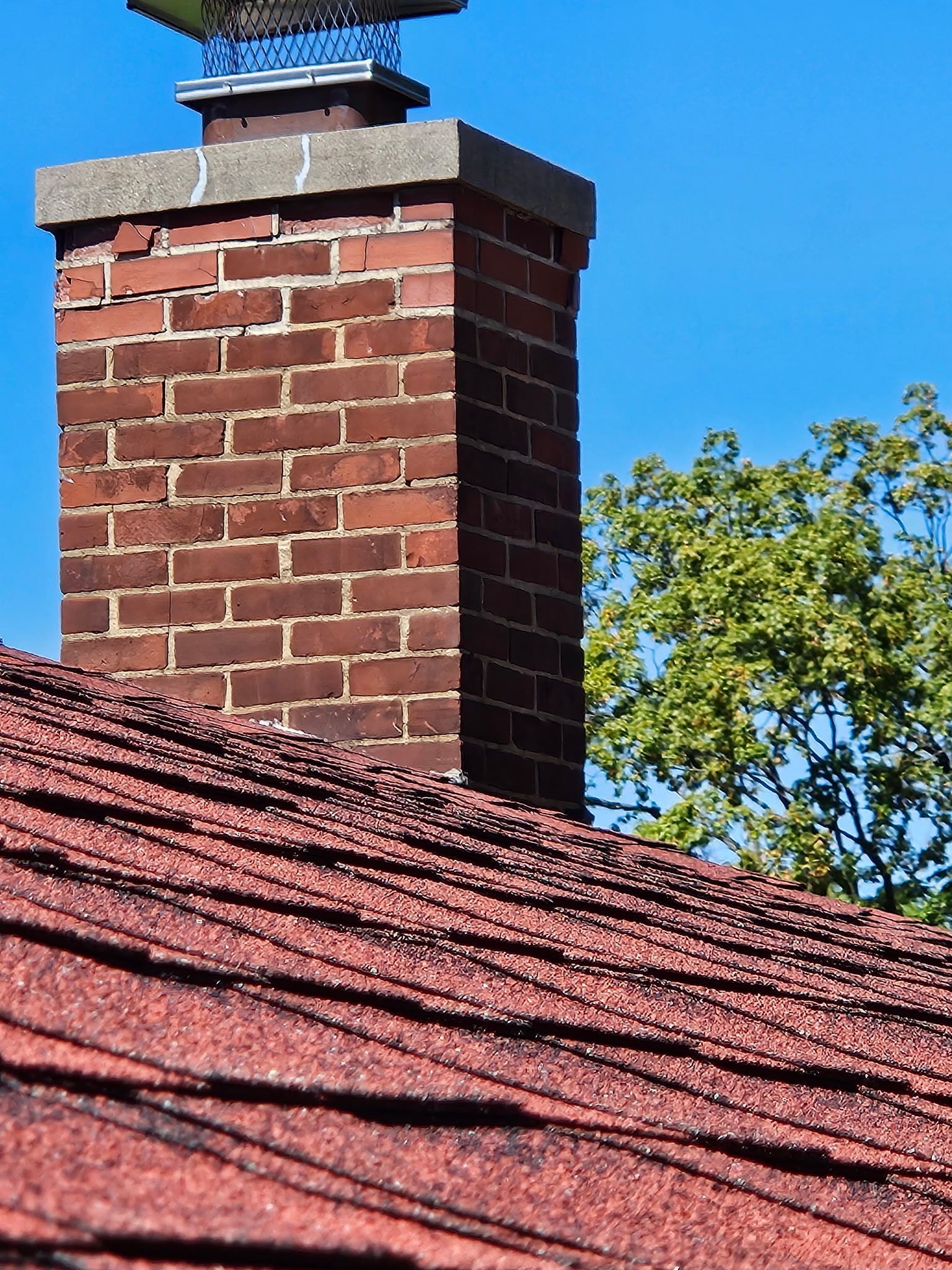 Brick chimney on a red-shingled roof against a bright blue sky, with a green tree in the background.