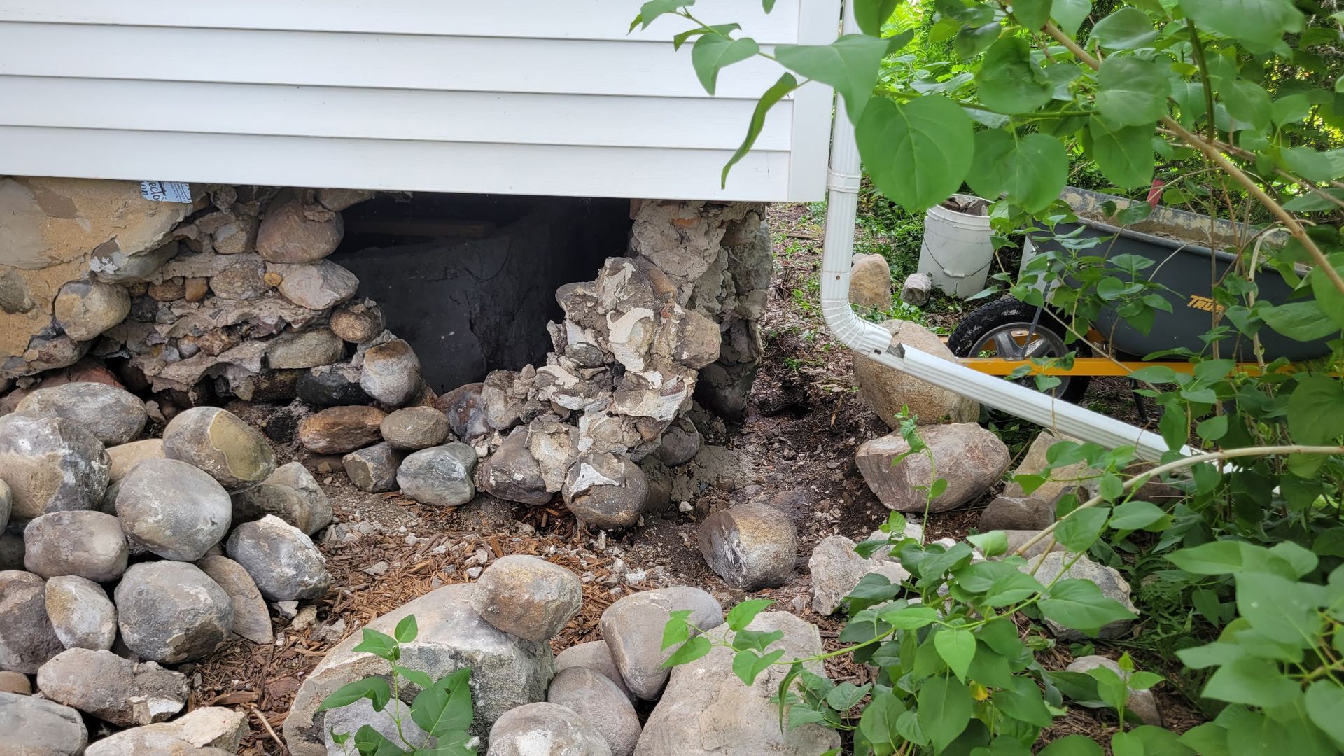 Damaged foundation of a white-sided building with exposed rocks and earth, surrounded by greenery.