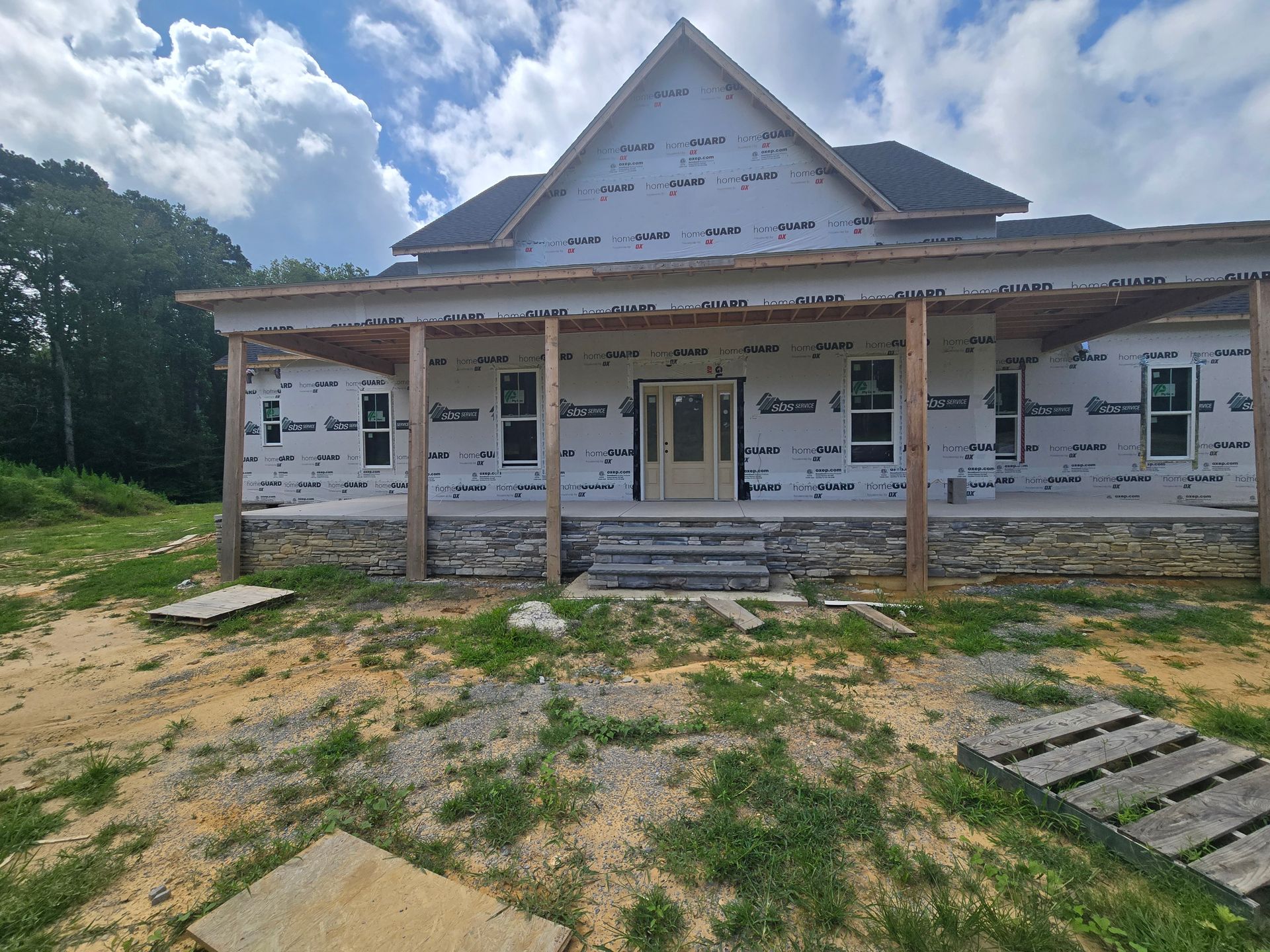 Partially built house with white siding and porch, set on a grassy lot under a cloudy sky