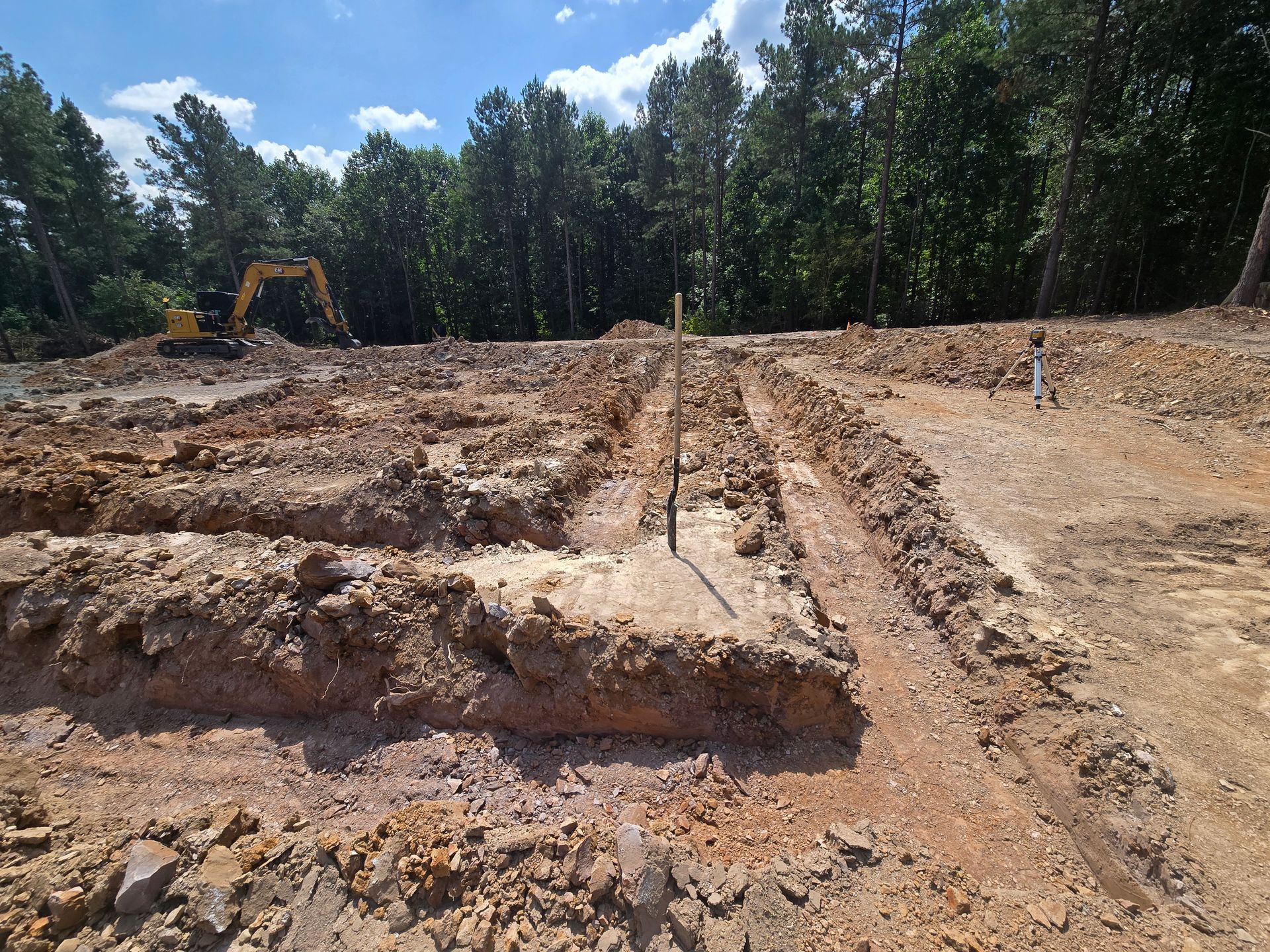 Excavation site with dirt trenches, an excavator, and trees under a blue sky.