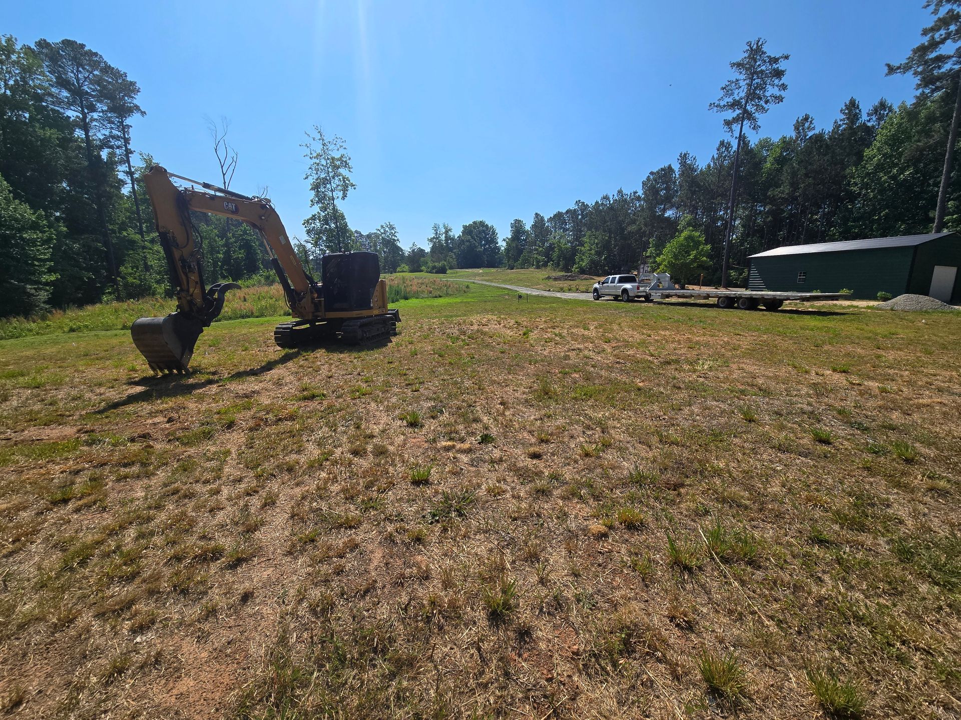 Excavator in a dry grassy field near trees and a small building under a clear blue sky