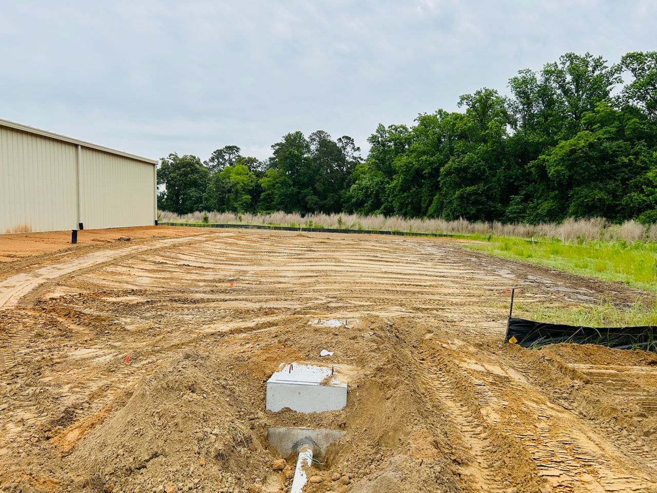Graded dirt lot beside a beige metal building and tree line, with a small concrete block in the foreground
