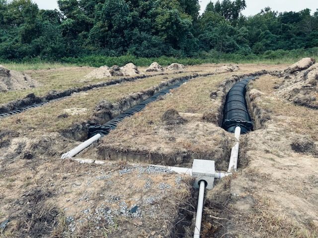 Plastic-lined drainage trenches through a grassy field, with pipes and dense trees in the background