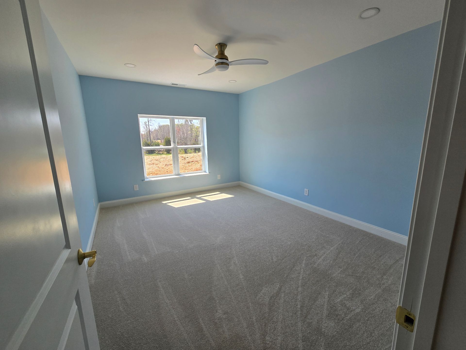 Empty blue bedroom with gray carpet, ceiling fan, and sunlight through a window.
