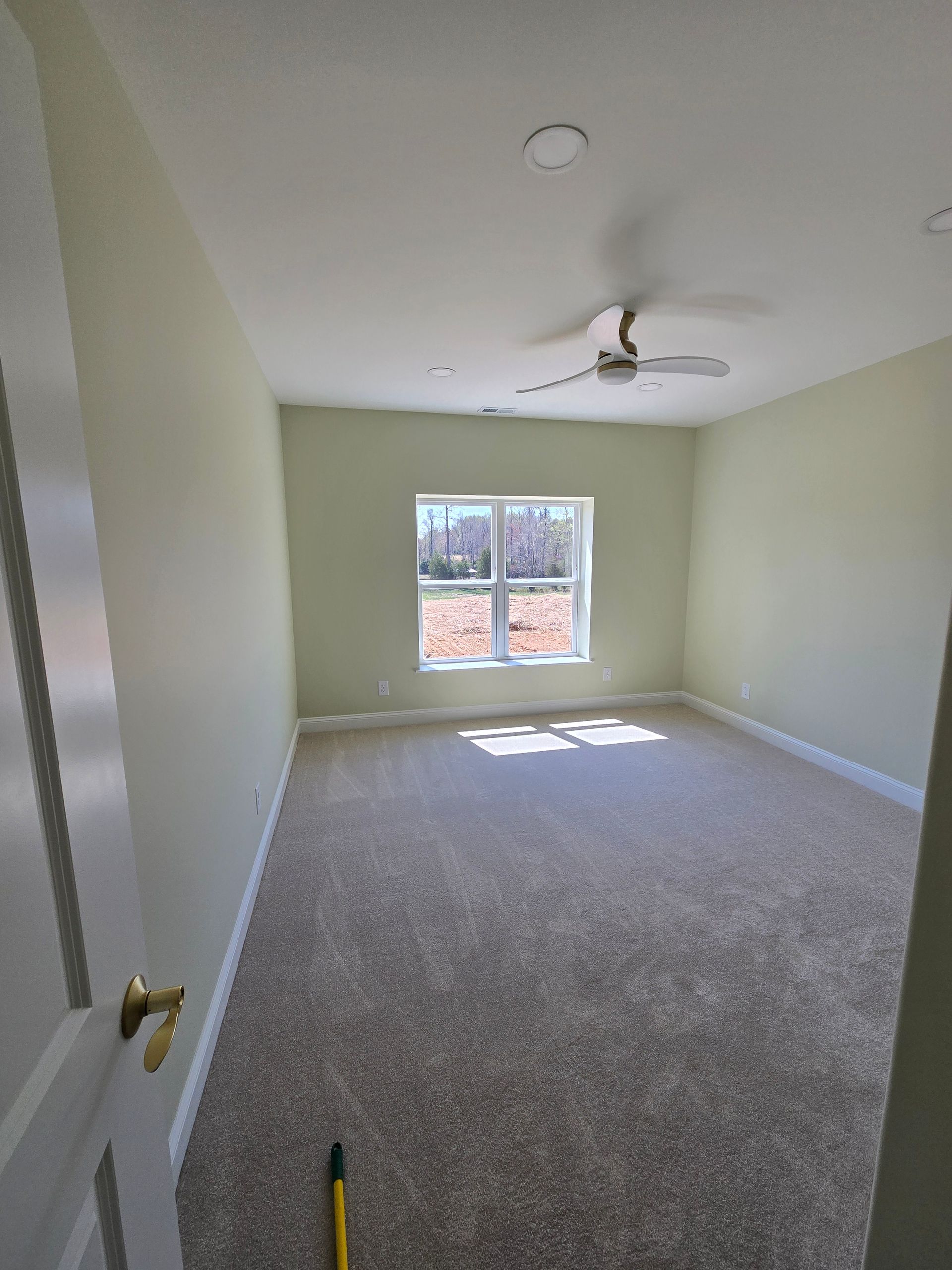 Empty carpeted bedroom with pale green walls, ceiling fan, and window letting in sunlight.