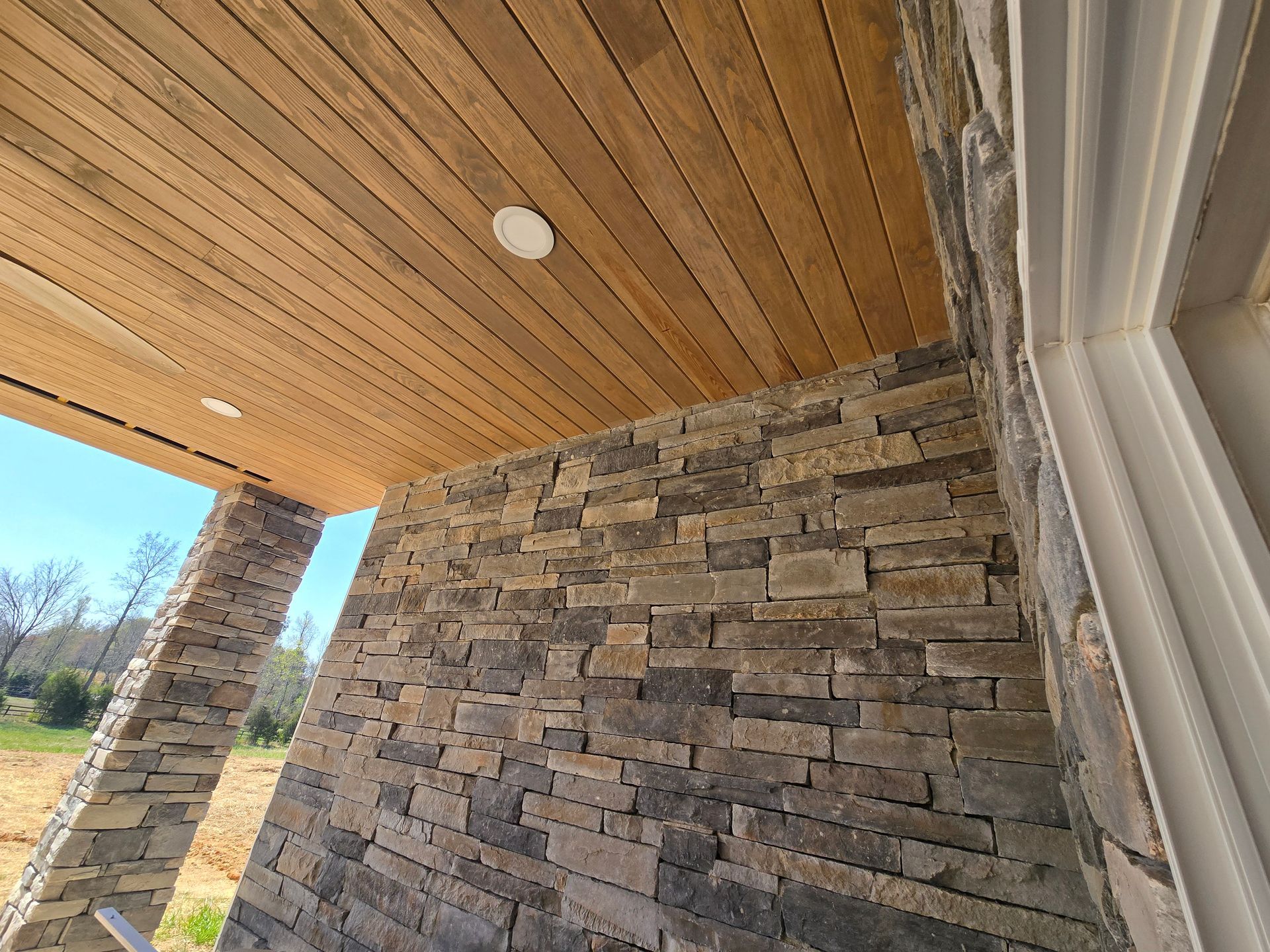 Covered porch with stone wall, wood ceiling, and windows on a sunny day