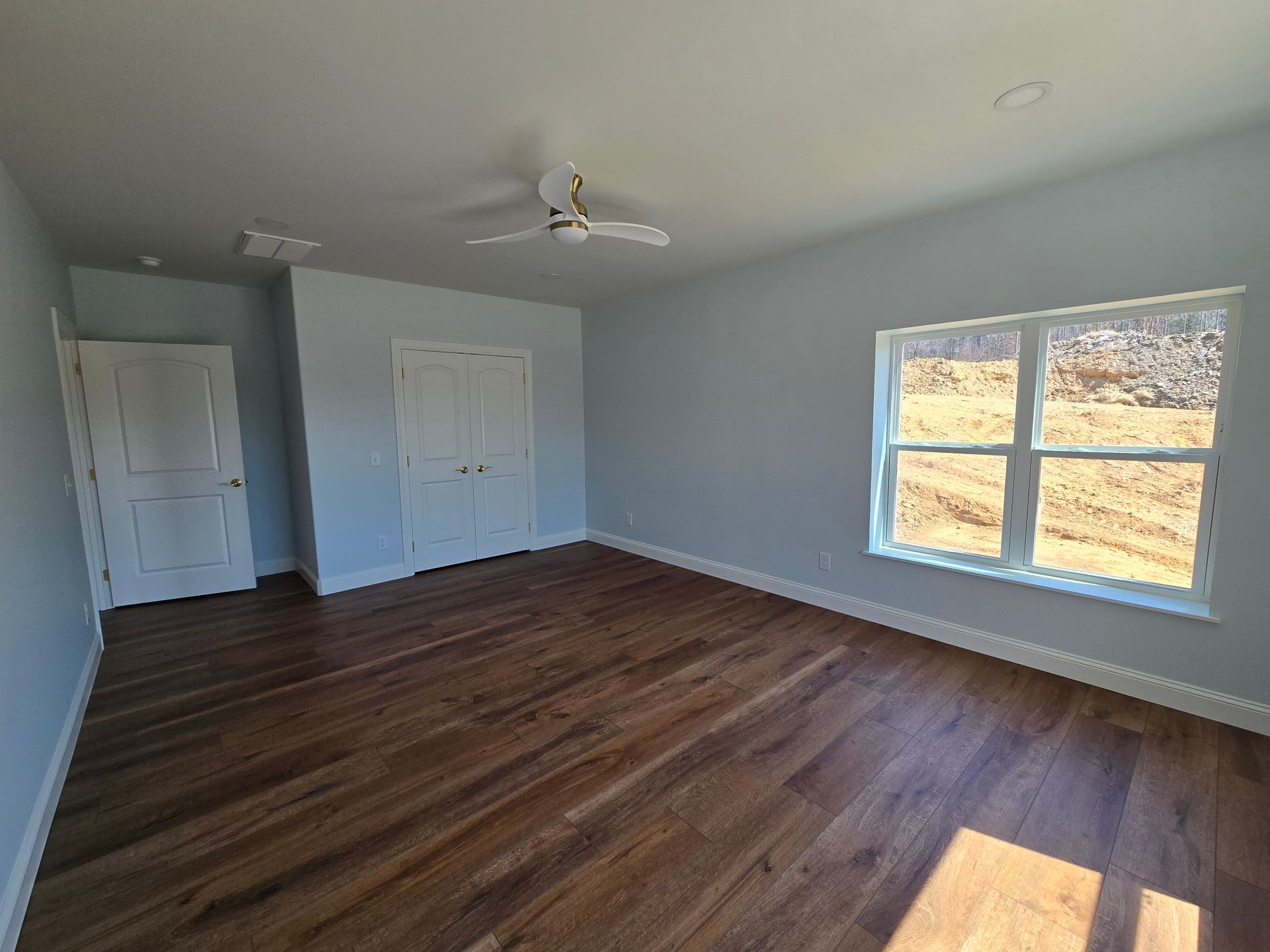 Empty bedroom with gray walls, dark wood floors, white doors, ceiling fan, and large sunlit window