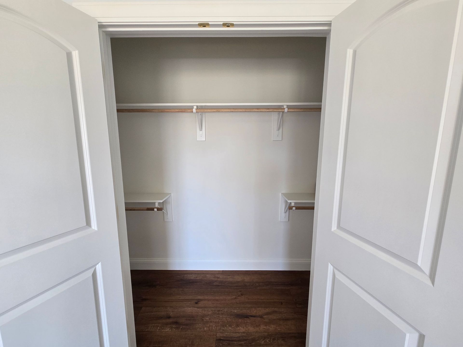 Open white closet with shelves, hanging rod, and dark wood floor