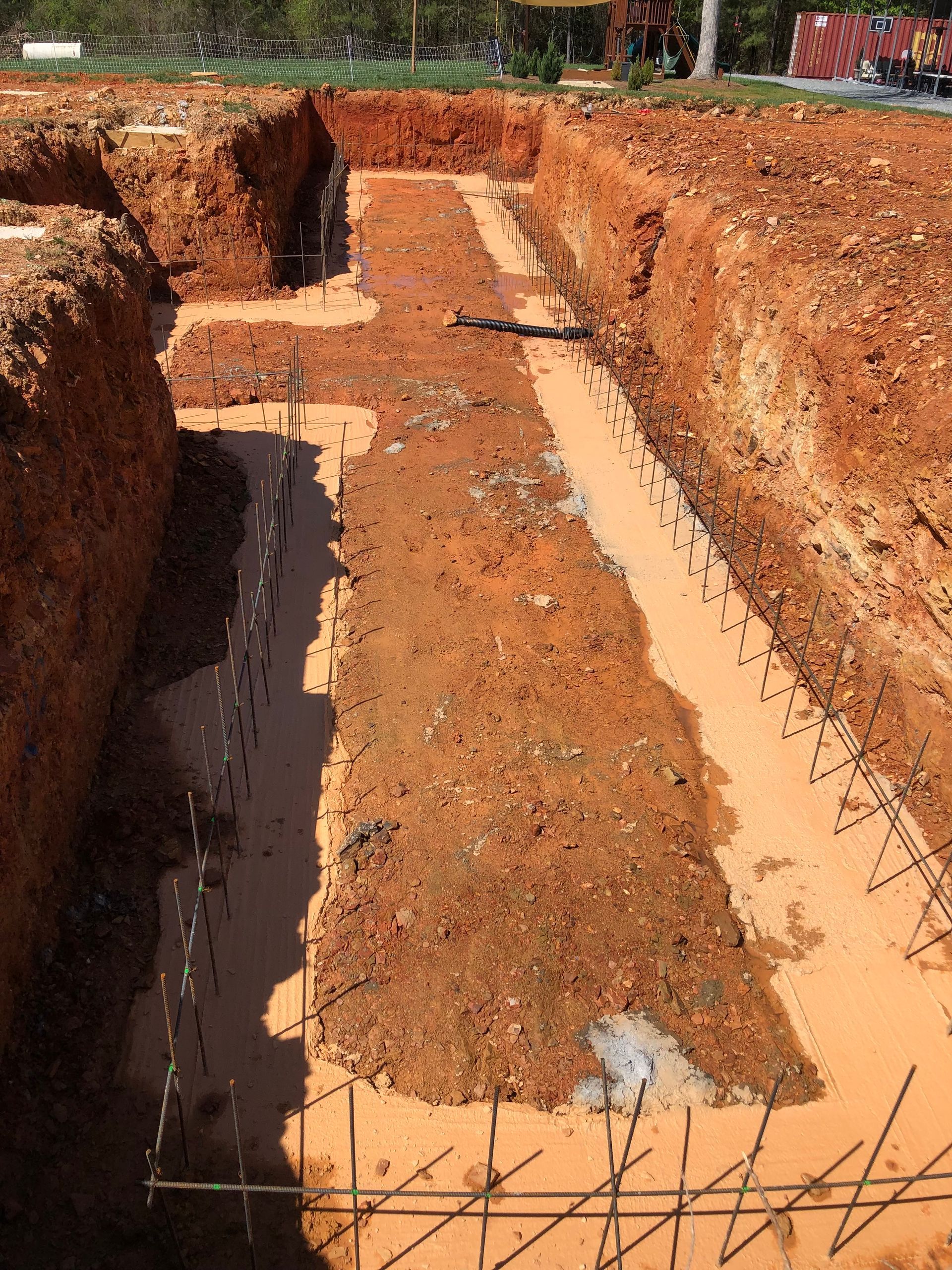 Excavated trench with red dirt and concrete footings, showing a building foundation under construction.