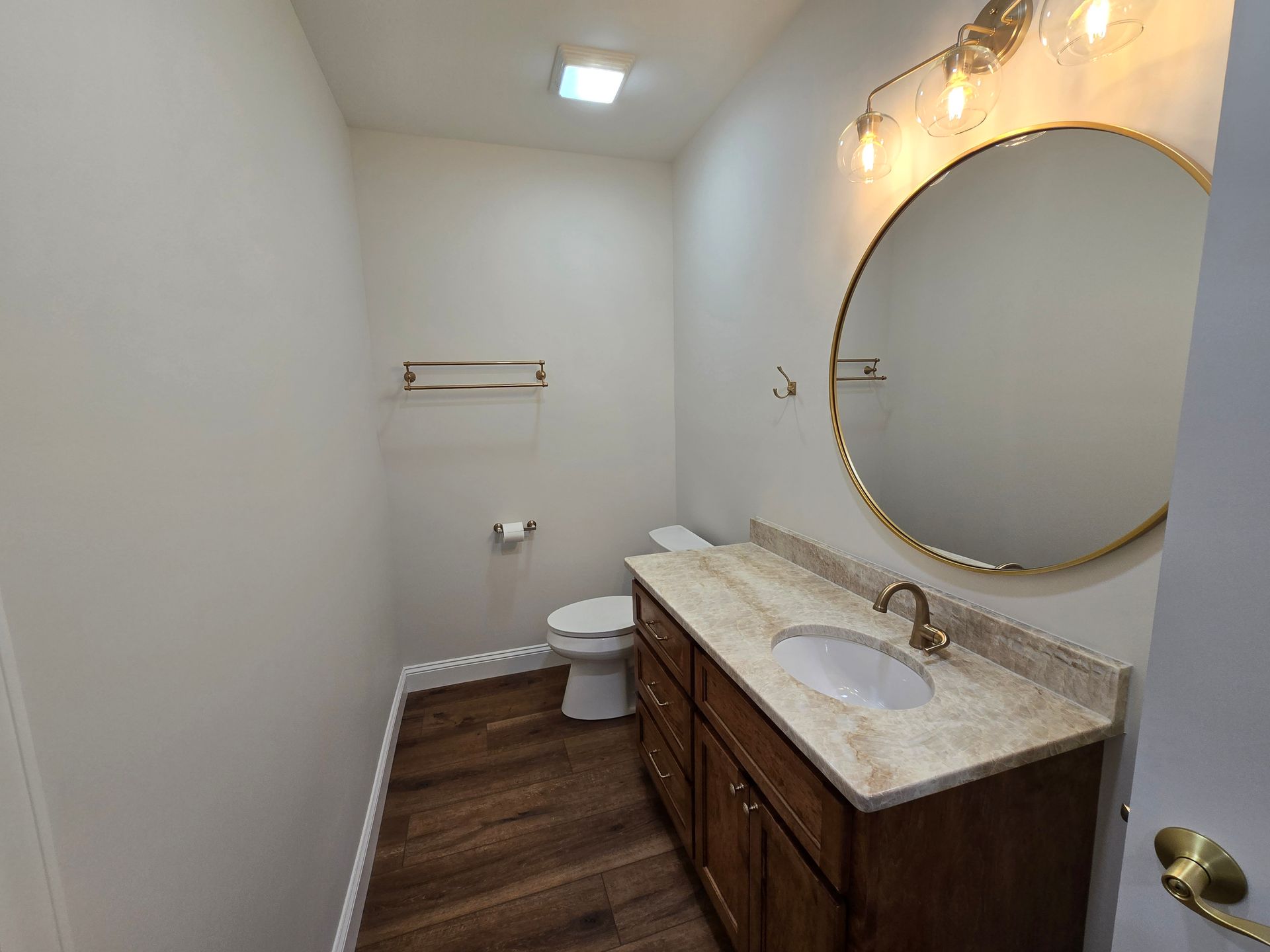 Narrow bathroom with double vanity, round mirror, toilet, and wood-look floor.