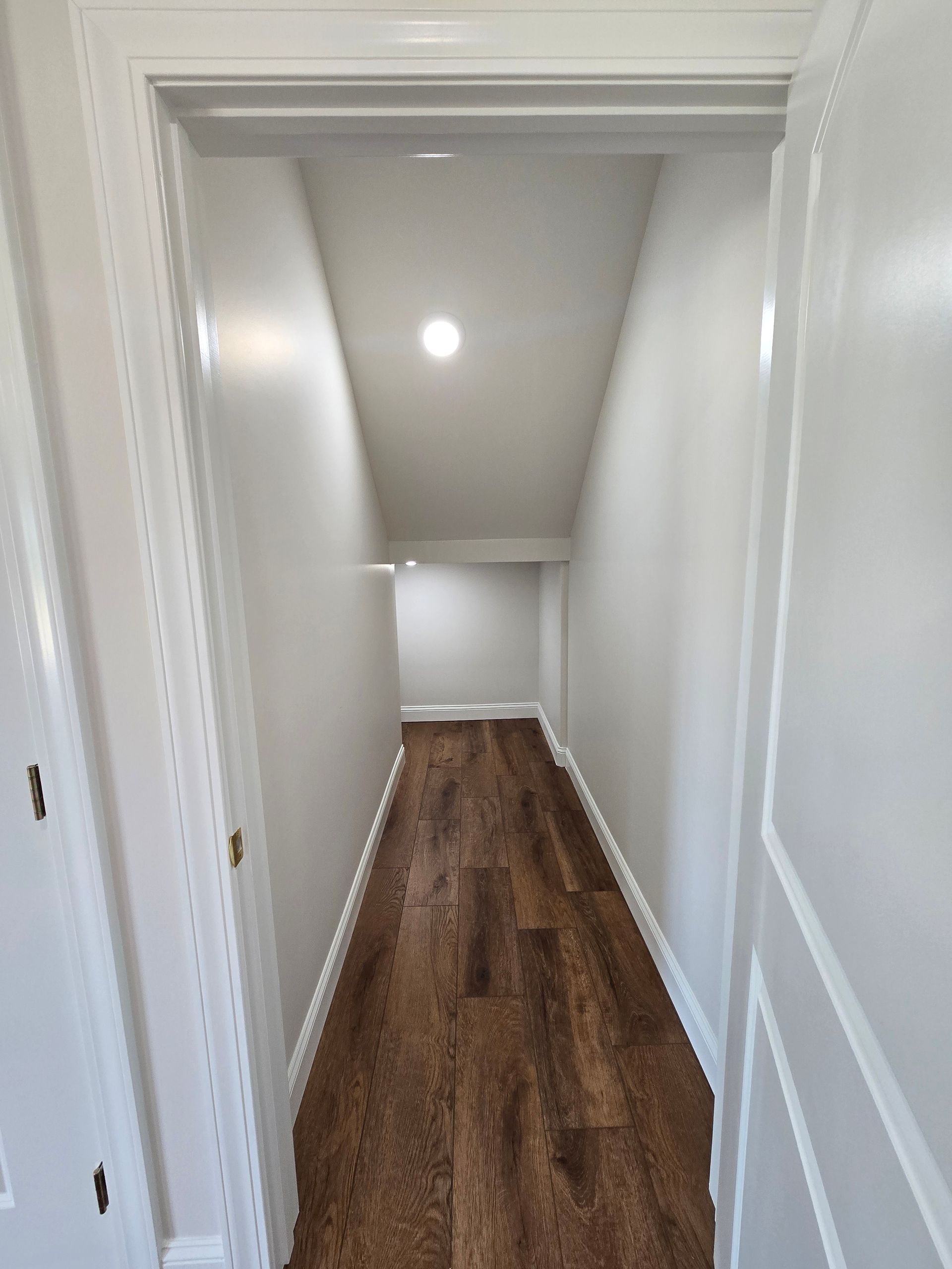Narrow white hallway with wood floor and recessed ceiling light, viewed from an open doorway