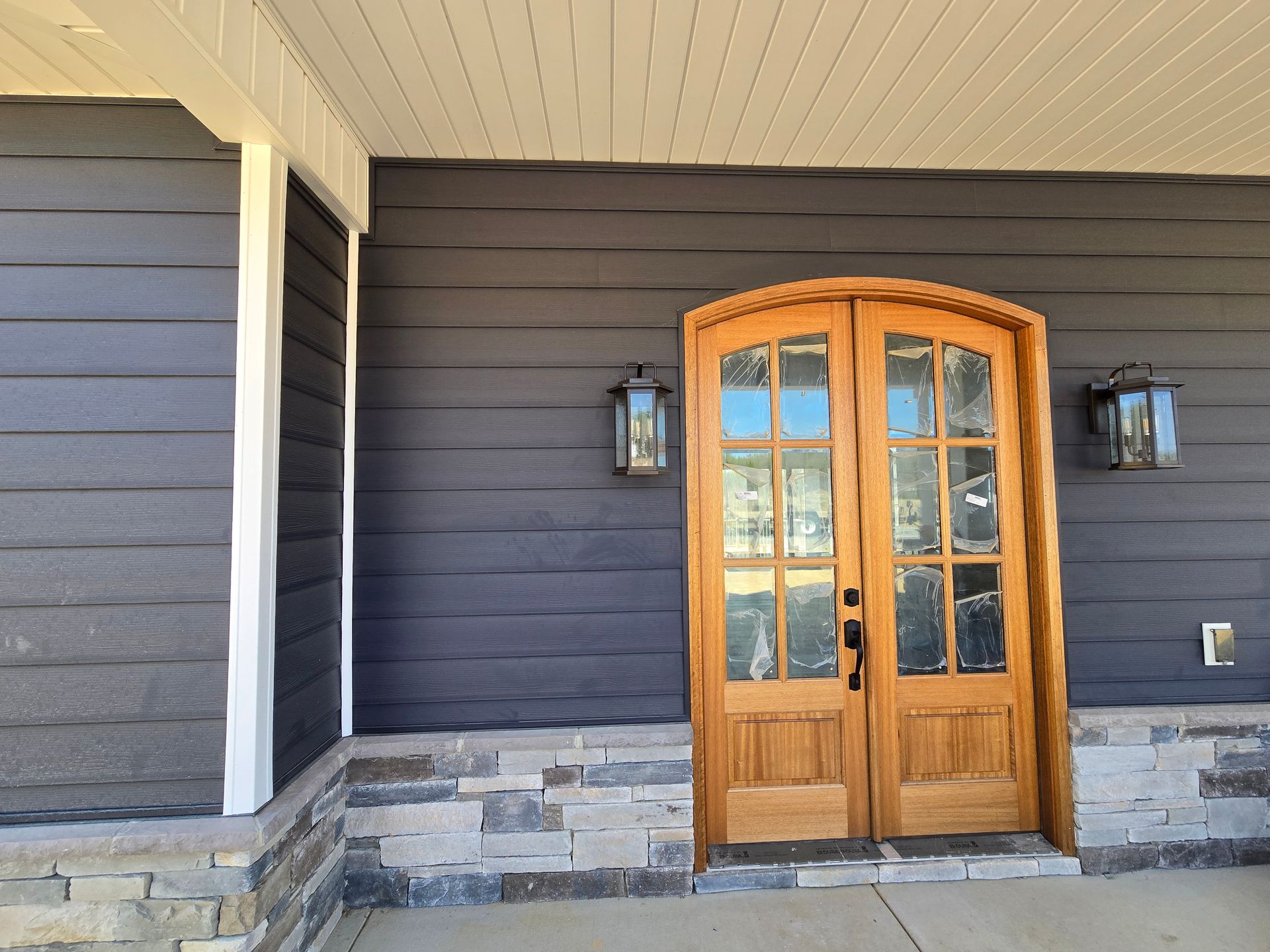 Front porch with wooden double doors, dark siding, and stone accents, flanked by two lanterns.